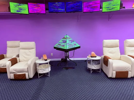 White recliners with salt lamps on side tables, the glowing pyramid between them and colorful screens along the top of the purple wall.